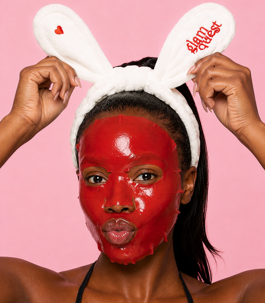 A woman with a glossy red face mask wears a white bunny headband with red embroidered text reading 'slam queen' against a solid pink background.